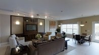 Located at 300 Northwoods Rd in Middletown, NY, this single-floor community space offers a welcoming and well-appointed gathering area designed for both relaxation and socializing. The open-concept layout seamlessly connects a lounge area with a dedicated dining space, creating a versatile environment for residents and guests alike.

The lounge is anchored by a rich, dark wood built-in entertainment center housing a flat-screen television, flanked by decorative shelving and accent lighting that adds warmth to the room. Track lighting runs across the ceiling, providing adjustable illumination throughout the space. Comfortable upholstered seating, including a generous sofa and coordinating armchairs in neutral tones, is arranged to encourage conversation and ease.

Large windows along the perimeter flood the space with natural light and offer views of the surrounding landscape, enhancing the open and airy feel. The dining area, positioned near the windows, features contemporary tables and chairs, making it ideal for casual meals or group activities. Light-toned tile flooring flows throughout, tying the spaces together cohesively. Decorative artwork and greenery add finishing touches that contribute to the inviting, modern ambiance of this community gathering space.