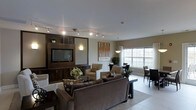 Located at 300 Northwoods Rd in Middletown, NY, this single-floor community space offers a welcoming and well-appointed gathering area designed for both relaxation and socializing. The open-concept layout seamlessly connects a lounge area with a dedicated dining space, creating a versatile environment for residents and guests alike.

The lounge is anchored by a rich, dark wood built-in entertainment center housing a flat-screen television, flanked by decorative shelving and accent lighting that adds warmth to the room. Track lighting runs across the ceiling, providing adjustable illumination throughout the space. Comfortable upholstered seating, including a generous sofa and coordinating armchairs in neutral tones, is arranged to encourage conversation and ease.

Large windows along the perimeter flood the space with natural light and offer views of the surrounding landscape, enhancing the open and airy feel. The dining area, positioned near the windows, features contemporary tables and chairs, making it ideal for casual meals or group activities. Light-toned tile flooring flows throughout, tying the spaces together cohesively. Decorative artwork and greenery add finishing touches that contribute to the inviting, modern ambiance of this community gathering space.