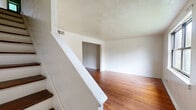 This two-story home at 99 Woodland Dr in Hartford, CT is efficiently laid out across two evenly divided floors. The main level features two living areas with warm hardwood floors, white walls, and a staircase that connects to the upper floor. Natural light filters in through windows offering views of surrounding greenery. The kitchen on the main level is equipped with wooden cabinetry, dark countertops, a stainless steel sink, and a window overlooking the outdoors, complemented by dark tile flooring. Upstairs, two bedrooms continue the hardwood floor aesthetic, each featuring windows that bring in natural light and tree-lined views. The primary bedroom is notably spacious, offering two windows and ample wall space. A hallway connects the upper-level rooms. The full bathroom features green ceramic tile walls surrounding a bathtub, with a window providing natural light and outdoor views. Textured ceilings are present throughout the home, contributing to its classic character. The layout is compact yet functional, making efficient use of its space across both levels.