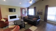 This single-floor home offers a welcoming and well-appointed living space designed with comfort and functionality in mind. The living room serves as the heart of the home, showcasing vaulted ceilings that lend an open, airy feel to the space. A white brick fireplace anchors one wall, providing a classic focal point and a cozy atmosphere. A ceiling fan is centrally mounted, ensuring year-round comfort. The room is finished with rich hardwood-style flooring that contrasts beautifully with the warm, earthy wall tones, including a distinctive accent wall in deep mauve that adds a touch of personality and depth. Natural light filters in through a window fitted with blinds and framed by dark curtains, brightening the interior. The living area is thoughtfully arranged with a large dark leather sofa, a sturdy rectangular coffee table, and a patterned area rug that ties the color palette together. A wall-mounted flat-screen television is positioned for optimal viewing. Decorative artwork and a lush indoor plant further enhance the space, adding life and character. The overall ambiance is warm, inviting, and tastefully styled, reflecting a comfortable and lived-in home.