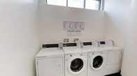 This shared laundry facility is a compact, single-floor space designed with functionality and cleanliness in mind. The room features bright white walls and is bathed in natural light streaming through a row of windows positioned near the ceiling, creating a fresh and airy atmosphere.

The facility is equipped with a lineup of commercial-grade Speed Queen washing machines and dryers, all finished in clean white. The machines are arranged side by side along the wall, providing multiple units for simultaneous use. Each unit features an easy-to-read digital control panel, and coin-operated mechanisms are in place for convenient operation.

Instructional signage is clearly posted on the wall above the machines, offering users guidance on proper machine operation and usage procedures. The overall layout is straightforward and user-friendly, maximizing the available floor space while keeping the room uncluttered.

The space is well-maintained, with a clean, minimalist aesthetic throughout. The combination of ample natural light, organized equipment placement, and clear informational displays makes this laundry room a practical and welcoming shared amenity.