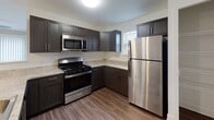 This single-floor residence offers a thoughtfully designed open-concept layout that seamlessly connects the kitchen and living areas. The kitchen is a standout feature, showcasing dark espresso cabinetry, granite-look countertops, and a full suite of stainless steel appliances including a gas range, built-in microwave, and refrigerator. A peninsula with an integrated sink and pendant lighting overhead bridges the kitchen to the living space, providing both functionality and visual appeal. The living area is generously proportioned, finished with plush neutral carpeting and warm beige walls. A ceiling fan anchors the room, while vertical blinds on a sliding glass door allow for controlled natural light and outdoor access. The flooring transitions from rich wood-look vinyl in the kitchen and entryway to soft carpet in the living and sleeping areas, creating a warm contrast throughout. The bedroom is bright and comfortable, with large windows dressed in horizontal blinds and its own ceiling fan for year-round comfort. The bathroom features wood-look vinyl flooring, a bathtub-shower combination, and a window that fills the space with natural light. Clean white walls and simple, modern fixtures give the bathroom a fresh, functional character. Overall, the space presents a clean, modern aesthetic well-suited for comfortable everyday living.