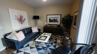 This single-floor space showcases a thoughtfully designed living area that balances bold style with refined comfort. The main living space is anchored by a sleek navy blue leather sofa accented with soft blush pink throw pillows, creating an appealing contrast that sets the tone for the room's modern aesthetic. A geometric-patterned area rug in navy, white, and gray ties the space together, adding visual energy underfoot.

A minimalist white coffee table with metallic legs serves as the centerpiece, styled with decorative trays and accessories that add a touch of sophistication. Opposite the seating area, a dark wood credenza provides ample storage while displaying carefully selected decor pieces and a framed landscape artwork that draws the eye. A lush fiddle leaf fig plant adds a natural, organic element that softens the room's contemporary lines.

Curated wall art, including a large botanical-inspired print, enhances the gallery-like atmosphere of the space. Warm, neutral-toned walls provide a versatile backdrop that allows the furnishings and decor to stand out. A sleek black floor lamp offers both functional and ambient lighting, completing this well-composed and inviting living environment that reflects a confident, modern design sensibility.