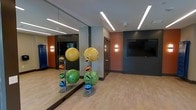 This single-floor fitness studio offers a well-appointed space designed for versatile exercise and group training activities. The room features full-length mirrored walls along one side, visually expanding the space and allowing users to monitor their form during workouts. The flooring is a smooth, light-toned hardwood-style surface, providing both durability and comfort underfoot.

Recessed linear LED lighting runs across the ceiling, delivering bright, even illumination throughout the studio. Accent wall sconces add a warm, secondary light source, complementing the overall modern aesthetic. The walls display a mix of neutral grays, warm terracotta, and deep charcoal tones, creating a dynamic and energizing atmosphere.

A wall-mounted flat-screen television is positioned prominently, ideal for streaming workout classes or displaying instructional content. Exercise ball racks hold a variety of stability and medicine balls in multiple sizes, catering to different fitness levels and training needs. A standard door provides access to adjacent areas of the facility. The overall design balances functionality with contemporary style, making it a practical and inviting environment for fitness enthusiasts.