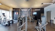 This fitness center is designed to provide a comprehensive workout environment within a single-level space. The room is anchored by a dramatic large-scale mural of a steel bridge spanning the full width of the back wall, rendered against a deep charcoal background that gives the space a sleek, modern aesthetic. The mural is reflected in a floor-to-ceiling mirror, visually expanding the room and adding depth. Pendant lighting and recessed ceiling fixtures provide bright, even illumination throughout. The facility is outfitted with a range of commercial-grade strength training machines and at least one stationary cycle, catering to varied fitness routines. Two wall-mounted flat-screen televisions are positioned on either side of the mural, offering entertainment during workouts. The flooring features durable rubberized material appropriate for a high-traffic exercise environment. Dark accent walls contrast with lighter ceiling tones, creating a focused and energetic atmosphere. The layout allows for comfortable movement between equipment, making the space both functional and visually engaging for users.