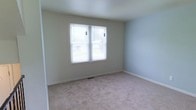 This two-floor home offers a clean, understated interior designed for comfortable everyday living. The image showcases a carpeted room with soft, neutral-toned walls in a light gray-blue palette, creating a calm and airy atmosphere. A centrally positioned double window fitted with horizontal blinds allows natural light to filter into the space, brightening the room and offering a view of the surrounding greenery outside. The light-colored carpet extends throughout the floor, adding warmth and a sense of continuity. White baseboards frame the room neatly, contributing to a tidy, well-maintained appearance. A floor vent is visible beneath the window, indicating a standard forced-air climate control system. The room's simple, open layout provides flexibility for various uses, whether as a bedroom, office, or sitting area. The overall aesthetic of the space is practical and move-in ready, with neutral finishes that provide a versatile backdrop for personalization. Spanning two floors, the home offers ample space for daily living across multiple levels.
