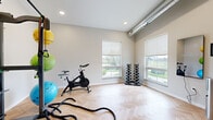 Located at 5532 38th St S in Fargo, North Dakota, this dedicated fitness studio is thoughtfully designed to maximize functionality within a compact, single-room layout. The space immediately impresses with its light-toned herringbone hardwood flooring, which lends a clean, modern aesthetic while providing a durable surface suited for a wide variety of workouts.

Natural light flows generously through multiple large windows fitted with neutral roller shades, offering views of the surrounding area while maintaining a bright and energizing atmosphere. An exposed industrial-style ventilation duct runs along the upper wall, adding an urban, contemporary character to the space.

The studio is well-equipped for diverse training modalities. A spin bike and battle ropes support cardio and conditioning workouts, while a full dumbbell rack with tiered weights accommodates strength training. A multi-functional Smart equipment rack holds resistance bands, yoga mats, medicine balls, and various accessories, making the space versatile for group or individual sessions. A wall-mounted Reflect interactive mirror provides guided workout programming, adding a tech-forward dimension to the studio experience.

Colorful stability balls complete the equipment offering, catering to balance and core training. A glass-paneled entry door with a modern wood frame provides a welcoming transition into the space. Recessed lighting complements the natural light, ensuring consistent illumination throughout.