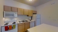 This single-floor space includes a well-organized kitchen that serves as a central feature of the unit. The kitchen is outfitted with light maple-toned wood cabinetry, offering generous upper and lower storage for pantry items and cookware. White appliances — including an electric coil range, built-in microwave, dishwasher, and full-size refrigerator — provide a complete set of everyday conveniences. A stainless steel double-basin sink sits beneath the upper cabinets, complemented by light-colored laminate countertops that extend across the full length of the cooking area. The layout follows a practical galley-style configuration, maximizing efficiency within the available space. A dining surface is partially visible in the foreground, suggesting a connected or open eating area just adjacent to the kitchen. The ceiling features a textured finish, and neutral-toned walls give the space a clean, understated appearance. Overhead lighting provides consistent illumination throughout the kitchen. Overall, the space presents a straightforward, functional design well-suited for daily living.