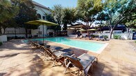 This outdoor amenity space is centered around a generously sized, crystal-clear swimming pool that serves as the focal point of the entire area. The expansive pool deck is finished in warm-toned stamped concrete, providing ample space for relaxation and recreation. Numerous reclining lounge chairs are arranged poolside, complemented by green market umbrellas that offer shaded respite on sunny days.

Mature trees are thoughtfully integrated throughout the space, providing natural shade and lending a lush, park-like atmosphere. A covered pergola structure anchors one end of the area, creating a dedicated shaded gathering zone equipped with additional seating. Landscape beds featuring ornamental shrubs, flowering plants, and decorative river rock borders line the perimeter, adding color and texture to the environment.

A separate patio area extends beyond the main pool deck, featuring picnic-style seating beneath umbrella shade and a decorative trellis overhead. This secondary outdoor space is connected via a flagstone-paved walkway, framed by well-maintained hedges and planters. Accent lighting from traditional post lanterns is positioned throughout, suggesting the space is equally enjoyable after dusk. The overall ambiance is resort-like, relaxed, and inviting, designed for both leisurely lounging and social gatherings.