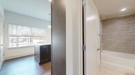 Located in the vibrant 75206 zip code of Dallas, Texas, this efficiently designed single-floor unit combines modern aesthetics with functional living. The open-concept kitchen and living area serves as the heart of the space, featuring rich dark espresso cabinetry, a generous granite-topped island with an undermount sink, and pendant lighting suspended from above. Stainless steel appliances, including a refrigerator, range, and dishwasher, complement the contemporary design. Wood-style flooring runs throughout, adding warmth and continuity across the space. A ceiling fan in the living area enhances comfort, while large windows fitted with roller shades invite generous natural light and frame views of mature trees and a private outdoor balcony. A glass-paned door provides direct access to the balcony, reinforcing the connection between interior and exterior. The bathroom is thoughtfully appointed with a deep soaking tub set within a full tile surround featuring a neutral travertine-style pattern, brushed nickel fixtures, and a built-in niche for added convenience. Clean white walls throughout the unit create a bright, airy ambiance that makes the space feel open and inviting.