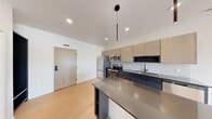 This contemporary single-floor residence showcases a thoughtfully designed open-concept layout with clean lines and a cohesive modern aesthetic. The kitchen serves as the centerpiece of the living area, featuring a large island with a dark quartz countertop, two-tone cabinetry in warm wood and deep charcoal tones, a classic tile backsplash, and a full suite of stainless steel appliances including a refrigerator, electric range, microwave, and dishwasher. A sleek pendant light hangs above the island, adding a stylish focal point. Light hardwood flooring flows consistently throughout the unit, enhancing the bright, airy atmosphere created by recessed lighting and crisp white walls. The bedrooms are generously proportioned, with floor-to-ceiling black-framed windows and sliding glass doors that open to an outdoor balcony, inviting natural light and outdoor views. A dedicated walk-in closet with wire shelving and an in-unit washer provides added convenience. The bathroom is appointed with modern matte black fixtures, an undermount sink set into a dark countertop, light-toned wood cabinetry, and large-format tile flooring. A sliding barn door detail adds a distinctive architectural touch to the interior hallway, completing this well-appointed and stylish space.