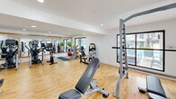 This fitness center in Phoenix, AZ, offers a bright and functional exercise space designed to accommodate a comprehensive workout experience. The single open-plan exercise room is finished with warm light wood flooring and crisp white walls, creating a clean, modern aesthetic complemented by recessed lighting throughout.

The cardio section is well-stocked with multiple Matrix treadmills, elliptical machines, and an Echelon spin bike, arranged in an organized row for ease of use. A wall-mounted television keeps users entertained during cardio sessions. Strength training is equally well-represented, with a multi-station cable machine, a squat rack, an adjustable weight bench, and a full dumbbell rack. Stability balls and additional free weights round out the functional fitness offerings.

Large framed mirrors line one wall, amplifying the sense of space and allowing users to monitor their form. Natural light pours in through multiple large windows and a sliding glass door, which opens directly to an outdoor courtyard and pool area — a standout feature that blurs the line between indoor and outdoor wellness. The layout flows intuitively, with cardio equipment on one side and strength and functional training zones on the other, making efficient use of the available space.