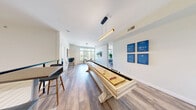 Located at 300 N Main St in Mansfield, Massachusetts, this single-floor community amenity space offers a well-rounded layout designed for both recreation and productivity. Warm hardwood-style flooring runs throughout, complemented by a bright, neutral color palette and contemporary artwork that lend the space a modern, welcoming ambiance.

The dual game rooms serve as a social centerpiece, featuring a full-length shuffleboard table beneath sleek pendant lighting, high-top bar seating, and a casual lounge area with a sectional sofa and street-view window. The open-concept kitchen and dining area is equipped with stainless steel appliances, white cabinetry, and a bold teal breakfast bar with pendant lights above — ideal for gatherings and events. An adjacent dining room offers a large conference-style table surrounded by cushioned chairs, floor-to-ceiling windows, and lush greenery that bring in abundant natural light and outdoor views.

A dedicated office area provides a quieter setting with small individual tables and patterned carpet, suited for focused work or study. The space also includes a closet for storage and a clean, well-appointed gender-neutral restroom with wood-tone flooring and a modern vanity. Together, these varied spaces create a versatile, amenity-rich environment for both social and professional use.