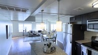 This single-floor residence showcases an open-concept layout that seamlessly integrates the kitchen, dining, and living areas into one cohesive, flowing space. The kitchen is appointed with granite countertops, dark cabinetry, and stainless steel appliances, including a full-size refrigerator. Two elegant pendant lights with cylindrical frosted shades hang above, providing warm, focused illumination over the central island or counter, which also serves as a casual gathering point. The dining area accommodates a glass-top table with upholstered chairs, bridging the kitchen and the living room. The living area features a neutral-toned sofa arrangement anchored by a low-profile area rug and coffee table, creating a comfortable and inviting atmosphere. Structural ceiling beams add architectural character throughout the space, while large windows along the perimeter allow generous amounts of natural light to fill the interior, contributing to an airy, open ambiance. The consistent use of cool tones, contemporary furnishings, and clean lines gives the space a modern, well-coordinated aesthetic throughout.