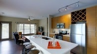 This single-story space showcases a thoughtfully designed open-concept layout that seamlessly connects the kitchen and living areas. The kitchen serves as the centerpiece, anchored by a generous white-topped island with an integrated sink and seating along one side, accommodating multiple bar stools. Warm honey-toned wood cabinetry extends to the ceiling, incorporating a distinctive diamond-pattern wine rack that adds both function and visual interest. Stainless steel appliances, including a gas range, built-in microwave, and a full-size refrigerator, complete the well-equipped cooking space. Track lighting mounted to the ceiling highlights the kitchen zone, while a ceiling fan with a light fixture serves the adjacent living area. The living space benefits from abundant natural light streaming through multiple windows dressed with patterned curtains, offering views of surrounding greenery. Dark hardwood-style flooring runs throughout, providing a grounding contrast to the light-colored surfaces. The overall ambiance is modern yet welcoming, combining functional design with clean lines and warm material tones that create a cohesive, comfortable atmosphere throughout the space.
