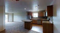 This single-story space showcases an open-concept layout where the kitchen flows naturally into the adjacent living area. The kitchen is well-appointed with warm medium-toned wood cabinetry that lines the perimeter walls, providing generous storage throughout. A slate-style tile backsplash adds texture and visual interest behind the sink and countertops. Stainless steel appliances, including a refrigerator, dishwasher, and microwave, lend a modern, functional quality to the space. A double sink is positioned beneath a window that allows natural light to brighten the work area. A central kitchen island with a light-toned countertop offers additional prep space and serves as a natural dividing point between the kitchen and the living zone. The kitchen features durable hardwood-style flooring, while the adjacent living area transitions to plush wall-to-wall carpeting, creating a comfortable and defined gathering space. Recessed lighting and a textured ceiling are consistent throughout, contributing to a cohesive and practical interior aesthetic. The overall layout is efficient and well-suited for everyday living.