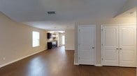 This single-story home offers a clean, open floor plan designed for comfortable everyday living. The main living area is spacious and welcoming, featuring rich, dark hardwood-style flooring that runs throughout the open space, creating a warm and cohesive aesthetic. Neutral beige walls and a textured ceiling complement the overall design, lending a bright and airy atmosphere to the interior. Natural light filters in through multiple windows dressed with white blinds, enhancing the sense of openness throughout the space. The living area flows seamlessly into the kitchen, which is equipped with dark-finished cabinetry and a pendant light fixture above, adding a subtle touch of elegance. A door at the far end of the kitchen area provides convenient access to the exterior. On one side of the main living space, two classic white panel doors with arched detailing lead to additional rooms, offering a neat and organized layout. Ceiling-mounted HVAC vents are integrated throughout, ensuring year-round climate comfort. The overall design of this home balances practicality with a timeless, understated style, making it a functional and inviting single-floor living environment.