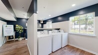 This shared laundry facility offers a clean, modern, and functional environment designed to accommodate multiple users with ease. The space is organized into distinct washing and drying zones, with several top-load washers and front-accessible dryers arranged in rows for convenient use. A bold two-tone color scheme — deep navy on the upper walls and crisp white below — gives the room a contemporary, polished aesthetic that sets it apart from a typical utility space. Warm wood-look flooring runs throughout, adding a touch of comfort underfoot and tying the design together cohesively. Recessed lighting provides even, well-distributed illumination across the room, while multiple windows along the walls invite abundant natural light and views of the surrounding greenery outdoors. A centrally positioned partial-wall partition subtly divides the space, creating separate functional areas without sacrificing openness. A wall-mounted payment kiosk provides convenient, cashless machine operation. Decorative framed artwork and potted greenery add thoughtful finishing touches, making the space feel welcoming rather than strictly utilitarian. Safety features including a fire extinguisher and clearly marked exit signage are also present. The overall layout is open and uncluttered, ensuring ease of movement and a pleasant laundry experience.