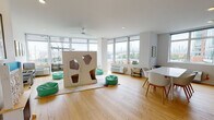 Located at 7601 River Rd in North Bergen, NJ, this single-floor facility is thoughtfully designed as a child-centered space that balances playful energy with calm, nurturing environments. The interior is finished with warm hardwood flooring throughout and crisp white walls that keep the atmosphere open and welcoming.

The main open area serves as the heart of the space, featuring floor-to-ceiling windows that flood the room with natural light and offer sweeping views of the surrounding landscape and distant skyline. A large freestanding play structure with cutout openings anchors the room, complemented by scattered bean bag seating in soft teal tones. A white dining table with modern chairs occupies one corner, providing a functional gathering area.

A dedicated learning room includes child-sized round tables and chairs, interactive activity boards mounted along the walls, and a comfortable glider chair — creating an ideal environment for structured play and early education. A full-length mirrored sliding closet adds depth and storage to this room.

Additional areas include a serene seating nook furnished with plush gray lounge chairs and contemporary artwork, as well as a quieter room adorned with a decorative macramé wall hanging and a colorful area rug. Each zone is thoughtfully appointed to support both active engagement and relaxed interaction for children and caregivers alike.
