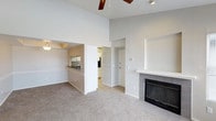 This single-floor residence is defined by its striking vaulted ceilings, which lend a sense of openness and airiness to the main living area. A ceiling fan is centrally positioned overhead, providing both practical ventilation and a visual anchor to the expansive ceiling height. The living room features neutral-toned carpeting and is anchored by a classic fireplace finished with dark tile surround and a clean white mantle, serving as a natural focal point within the space.

The open layout allows for a seamless visual connection between the living area and the kitchen, which is partially separated by a half-wall breakfast bar. The kitchen area features tile flooring, overhead cabinetry, and a sink positioned at the bar, offering both functionality and accessibility. A hallway visible from the living space leads toward additional rooms and a front entry door, suggesting a well-organized, efficient floor plan.

The overall color palette is light and neutral throughout, with white walls and soft gray tones contributing to a clean, understated aesthetic. Recessed ceiling detail in the kitchen area adds subtle architectural interest, distinguishing it from the vaulted main living space.