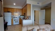 This single-floor residence offers a practical and inviting layout designed for comfortable everyday living. The kitchen is a standout feature, showcasing warm honey-toned wood cabinetry that extends to the upper walls, providing generous storage space. Equipped with a freestanding range, built-in microwave, and a countertop peninsula that serves as both a prep area and casual dining surface, the kitchen is both functional and well-organized. Light tile flooring runs throughout the space, lending a clean, cohesive aesthetic that reflects natural and ambient light effectively. The kitchen flows seamlessly into the living area, where a cushioned sofa anchors the space and contributes to the warm, relaxed atmosphere. A structural column defines the boundary between the two areas while maintaining an open feel. Recessed lighting in the kitchen ceiling ensures the workspace is well-lit, while HVAC vents are thoughtfully integrated into the ceiling throughout. A hallway visible from the main living area leads to additional rooms, hinting at further living and bathroom spaces beyond. The overall ambiance is cozy and residential, with a neutral color palette of creams and warm browns creating a welcoming environment throughout.