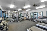 Located at 30300 Antelope Rd in Menifee, CA, this fitness center is a fully equipped exercise facility designed to support a well-rounded workout routine. The room features dark, durable carpeted flooring and a textured ceiling outfitted with recessed fluorescent lighting and ceiling fans to maintain a comfortable environment during workouts.

The space is well-stocked with a diverse range of equipment. Cardio offerings include multiple treadmills and upright stationary bikes, providing options for various aerobic exercises. A multi-station cable machine serves as the centerpiece for strength training, complemented by a dedicated dumbbell rack stocked with a range of free weights and a flat workout bench. Cable attachments and resistance accessories are neatly organized along the wall.

Large windows along two walls allow abundant natural light to fill the room while offering views of the surrounding outdoor greenery, creating an open and energizing atmosphere. Full-length mirrors on one wall visually expand the space and allow users to monitor their form during exercise. Wall-mounted televisions provide entertainment, and informational fitness charts are posted for user reference.

Multiple entry points, including a glass door leading to an adjacent lounge area and an exterior door for direct outdoor access, enhance the functionality and flow of this practical, community-oriented fitness facility.