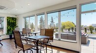Located on California Avenue SW in Seattle's vibrant West Seattle neighborhood, this rooftop amenity space offers a well-appointed indoor-outdoor retreat designed for gathering and relaxation. The interior lounge area is bright and contemporary, featuring light hardwood-style flooring, recessed lighting, and a wall of large floor-to-ceiling windows that flood the space with natural light. High-top bistro tables and chairs are arranged throughout, providing a casual setting with direct sightlines to the surrounding treeline and open sky. A living green wall adds a natural accent, while a clean, accessible restroom with backlit mirror and tiled finishes completes the interior. The expansive wraparound rooftop terrace is a standout feature, finished with large-format pavers and enclosed by a bold red railing. The outdoor area is outfitted with a generous sectional seating arrangement surrounding a central fire pit, Adirondack chairs, raised planter beds, and additional bistro seating. Overhead string lights create an inviting ambiance for evening use. Panoramic views of Seattle's lush canopy, neighboring rooftops, and distant skyline are visible in multiple directions, making this rooftop terrace an exceptional amenity for communal enjoyment year-round.