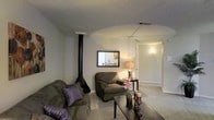 This single-floor space offers a comfortable and functional layout designed for everyday living. The living area serves as the heart of the home, featuring a standout black freestanding wood-burning stove with a sleek vertical pipe that draws the eye upward toward the textured popcorn ceiling. The room is generously proportioned, providing ample space for seating arrangements, with plush upholstered furniture and a dark wood coffee table lending a warm, casual ambiance. Neutral-toned walls and light carpeting create a soft, cohesive backdrop throughout. A framed mirror on the wall adds depth to the space, while a decorative table lamp and accent flowers introduce touches of warmth and personality. An arched ceiling detail subtly separates the main living area from a connecting hallway, which leads further into the home and is softly illuminated by a wall-mounted light. HVAC vents are integrated into the ceiling, ensuring year-round comfort. The overall atmosphere is relaxed and welcoming, with a blend of practical features and homey details that make this a comfortable, move-in-ready living environment.