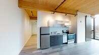 Located at 7373 N Philadelphia Ave in Portland's St. Johns neighborhood, this thoughtfully designed single-floor unit showcases a compelling blend of modern aesthetics and warm natural materials. The space is immediately distinguished by its striking tongue-and-groove wood ceiling, which runs throughout the open layout and adds a rich, organic character to the interior. Industrial-style track lighting mounted on black rails complements the natural ceiling tones while providing ample illumination.

The kitchen is efficiently designed with charcoal gray lower cabinetry, white upper shelving, and a full suite of stainless steel appliances, including a refrigerator, electric range, and dishwasher, all set against a clean light countertop and understated backsplash. Light wood-toned luxury vinyl plank flooring flows seamlessly throughout the living areas, tying the space together with a cohesive, contemporary feel.

The main living area is bathed in natural light through large, floor-to-ceiling windows framed by exposed wood structural columns, offering open views of the surrounding Portland neighborhood. A wall-mounted mini-split unit provides climate control. The bathroom features a clean, minimalist design with dark vanity cabinetry, a white countertop, a large mirror, and a full tub-shower combination. An in-unit washer is also present, adding everyday convenience to this well-appointed space.