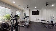 Located at 5750 Curtis Clark Dr in Corpus Christi, TX, this fitness center is a thoughtfully designed, single-floor facility built to support a comprehensive workout routine. The space features crisp white walls accented by crown molding, dark rubber flooring for durability and comfort underfoot, and recessed lighting complemented by sleek black ceiling fans that maintain airflow throughout the room.

The cardio zone is outfitted with multiple treadmills, an elliptical trainer, and a stationary bike, positioned to take advantage of a bank of large windows that allow abundant natural light and views of the outdoor pool and lush landscaping. A wall-mounted flat-screen television adds entertainment options during cardio sessions.

The strength training area offers a diverse selection of equipment, including a multi-station cable and weight machine, a full dumbbell rack with an integrated mirror wall that visually expands the space and supports proper form, and multiple weight benches. Additional free weights and a squat/weight rack round out the strength offerings.

Access to the fitness center is provided through elegant French doors, reinforcing the polished, upscale aesthetic of the facility. The combination of natural light, ample equipment variety, and clean architectural finishes makes this fitness center a functional and inviting space for users of all fitness levels.