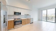 Located on the penthouse level at 999 NW Seventh St in Miami's vibrant urban core, this one-bedroom, one-bathroom residence offers a sleek, contemporary design throughout. The open-concept layout is anchored by a well-appointed kitchen featuring warm wood lower cabinetry, crisp white upper cabinets, a neutral tile backsplash, stainless steel appliances including a range, microwave, dishwasher, and French door refrigerator, all set against light large-format tile flooring that runs throughout the unit. Soaring ceilings in the kitchen and living area enhance the sense of openness and airiness. The bedroom is generously proportioned, bathed in natural light from floor-to-ceiling windows framing impressive views of the Miami skyline and surrounding cityscape. A private balcony extends the living space outdoors, offering additional city views. The bathroom is finished with a wood-toned vanity, white quartz countertop, large-format mirror, and a glass-enclosed shower. A spacious walk-in closet with built-in wire shelving provides ample storage. A hallway connects the rooms cohesively, completing this clean, modern residence ideal for urban living in Miami.