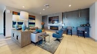 This single-floor amenity space is thoughtfully designed to offer a dynamic blend of comfort, function, and modern style. The lounge area serves as a central gathering point, anchored by a sleek linear electric fireplace framed in textured gray tile and topped with a wall-mounted flat-screen television. Generous sectional and accent seating in neutral tones are accented with teal and blue throw pillows, complemented by a layered area rug and bold abstract artwork on the walls, creating an inviting and visually engaging atmosphere.

Adjacent to the lounge is a well-appointed kitchen and bar area, featuring a mosaic stone backsplash, under-cabinet accent lighting, stainless steel appliances including a full-size refrigerator, and bar-height seating. A distinctive bistro-style table with a natural wood stump base and white surface adds a unique organic touch to the space, perfect for casual dining or conversation.

The layout, as seen in the dollhouse overview, also reveals a billiards table and dedicated workspace seating, making this a truly multi-purpose environment. Large-format tile flooring flows seamlessly throughout, tying all zones together. Recessed lighting throughout ensures a bright, well-lit experience across every area of this versatile communal space.