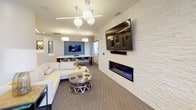 This well-appointed club room offers a thoughtfully designed communal space spread across a single floor, combining comfort and functionality in a contemporary setting. The lounge area serves as the centerpiece, featuring a sleek linear fireplace set into a striking white stacked-stone accent wall, above which a large flat-screen TV is mounted. Plush cream sectional sofas and a metallic coffee table create an inviting gathering space, while modern globe pendant lighting and recessed linear LED fixtures illuminate the room with a warm, sophisticated glow. Neutral-toned carpeting and clean architectural lines contribute to the space's polished aesthetic. Adjacent to the lounge, a dedicated dining area provides additional seating for groups, complemented by a wall-mounted display screen—ideal for presentations or entertainment. The fully equipped kitchen features warm wood cabinetry, a white quartz island with contemporary bar seating, stainless steel appliances including a full-size refrigerator, and a stone tile backsplash. A built-in coffee and beverage station adds a hospitality-forward touch. Large windows along the perimeter allow natural light to fill the space, offering views to the exterior streetscape. The overall ambiance is modern and refined, making this club room a versatile amenity suited for both relaxation and social gatherings.