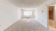 Located at 2445 Lyttonsville Rd in Silver Spring, Maryland, this single-floor unit offers a clean, straightforward layout designed for comfortable everyday living. The living area is generously proportioned, featuring wall-to-wall neutral carpeting and crisp white walls that create a bright, open atmosphere. A large window fitted with vertical blinds allows natural light to filter in, enhancing the sense of space throughout the room. Adjacent to the living area, the kitchen is efficiently designed with warm-toned wood cabinetry, granite-style laminate countertops, a stainless steel sink, and white appliances including a gas range and full-size refrigerator. The light flooring in the kitchen provides a clean contrast to the cabinetry and keeps the space feeling fresh. The bedroom is equally well-lit, with the same soft carpeting underfoot and neutral walls that offer a calm, restful environment. The bathroom features full tile surround in a classic grid pattern, a soaking tub with shower, chrome fixtures, and coordinating tile flooring, lending a timeless and functional finish. Overall, the unit presents a practical and well-maintained living space in a convenient Silver Spring location.