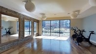 This single-floor fitness and yoga studio offers a welcoming, light-filled environment designed to support active pursuits and mindful practice. The space is anchored by a wall of floor-to-ceiling windows that flood the room with natural light and frame expansive views of a lush, landscaped courtyard garden. The rich hardwood flooring throughout provides a warm, polished surface well-suited for yoga, stretching, or group fitness activities. A striking stacked stone accent wall with framed openings adds a natural, earthy texture that contrasts beautifully with the clean white ceiling, lending the space a balanced, contemporary aesthetic. Overhead, large globe pendant lights provide soft, ambient illumination that complements the abundant natural light. Recessed ceiling speakers and ventilation fixtures suggest the space is thoughtfully equipped for structured classes and extended workouts. Exercise equipment, including a stationary bike, is visible near the windows, indicating the room's versatility for both cardio and mind-body disciplines. The overall ambiance is open, airy, and serene, with the combination of natural materials, generous glazing, and garden views creating an inviting atmosphere that encourages wellness and movement.