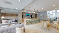 This single-floor communal space is designed with a clean, contemporary aesthetic that blends comfort and functionality. The open-plan layout centers around a sleek bar-height counter finished in light wood tones, lined with vibrant teal bar stools that add a pop of color against the neutral palette. Elegant cluster pendant lights hang above the counter, complementing the recessed ceiling lighting that keeps the space bright and inviting.

A wall-mounted flat-screen TV serves as a focal point behind the counter, set against a textured tile accent wall that adds visual depth and dimension. The counter area appears to double as a refreshment station, making it a natural gathering point for socializing.

The lounge seating area features plush gray sofas with decorative throw pillows, arranged on a patterned area rug over light hardwood-style flooring that runs throughout the space. Rattan accent chairs add a touch of organic warmth. Soft blue-gray walls contribute to the overall calm, airy ambiance.

A built-in mailbox unit is visible toward the rear, suggesting this is a residential amenity or lobby lounge space. Tasteful framed artwork and potted greenery complete the polished, welcoming environment.