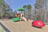 Located in Columbus, Georgia (31904), this single-level outdoor community space offers a welcoming environment surrounded by natural beauty. The grounds are characterized by gently sloping grassy areas that transition into a backdrop of tall pines and mixed deciduous trees, providing a natural buffer and a sense of privacy throughout the property.

A standout feature of this space is the dedicated playground area, equipped with a multi-level play structure complete with slides, a covered tower, and interactive elements, all set within a wood chip safety surface bordered by timber edging. This makes the area well-suited for families with children.

Concrete walkways wind through the property, providing smooth and accessible pathways connecting the various areas of the community. The well-maintained grounds include open lawn spaces that offer room for outdoor activities and relaxation. Neighboring residential buildings are visible in the background, reinforcing the community-oriented character of the space.

The mature tree line along the perimeter not only adds to the aesthetic appeal but also provides natural shade and a serene, park-like ambiance. Under clear blue skies, the combination of open green space, wooded edges, and community amenities creates an inviting outdoor environment for residents of this Columbus, GA neighborhood.