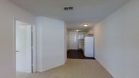 This single-story unit offers a straightforward, open-concept layout that connects the main living area to the kitchen in one continuous sight line. The living space features neutral-toned carpet, white walls, and a textured ceiling with a ceiling-mounted vent, contributing to a clean, functional aesthetic. A partial wall divides the entryway from the main living area, adding a subtle architectural detail while maintaining an open feel. The kitchen is positioned at the rear of the unit and is outfitted with white upper cabinetry, a white refrigerator, and a range, all set against dark hardwood-style flooring that contrasts with the carpeted living space. Flush-mount ceiling fixtures provide consistent overhead lighting throughout. A white paneled door at the far end of the kitchen likely leads to additional storage or a utility area. The overall design is practical and neutral, making the space adaptable for a variety of furnishing styles. The consistent use of white tones throughout creates a bright, cohesive atmosphere across the unit.