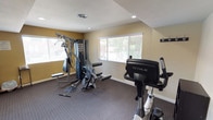 This fitness center is a thoughtfully designed, single-floor workout facility that offers a comprehensive range of exercise equipment within a comfortable and functional environment. The space is characterized by warm gold and cream-toned walls accented by bold deep red crown-level color bands, creating a motivating and energetic atmosphere throughout.

The main workout area is generously proportioned, featuring durable patterned carpet flooring that provides stability and comfort underfoot. A full-length mirrored wall spans one side of the room, visually expanding the space while allowing users to monitor their form during exercise. Natural light filters in through multiple windows fitted with horizontal blinds, offering outdoor views of surrounding greenery and keeping the space bright and inviting.

Equipment includes a multi-station cable and weight resistance machine suitable for a wide variety of strength training exercises, along with TRUE brand upright and recumbent cardio bikes. A dedicated alcove area provides additional space for equipment or stretching. Recessed ceiling lighting supplements natural light throughout.

A small entry or transition area includes a wall-mounted coat hook rack, a compact storage unit, and a window overlooking the exterior, offering a convenient space to prepare before or after a workout. The overall layout is practical and well-organized, making this fitness center a fully functional amenity space.