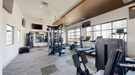 Located at 5555 W Ina Road in Marana, Arizona, this fitness center offers a single, spacious exercise room thoughtfully designed to support a wide range of workout styles and fitness goals. The room features soaring ceilings with a distinctive wood-paneled tray accent, lending a modern and polished aesthetic to the space. Crisp white walls are paired with a patterned carpet in muted green and grey tones, creating a clean and professional environment.

Natural light flows in through large, glass garage-style doors and tall windows along the perimeter, providing views of the surrounding outdoor areas. Multiple wall-mounted flat-screen televisions are positioned throughout the room for entertainment during workouts.

The facility is comprehensively equipped with a wide array of commercial-grade machines, including treadmills, ellipticals, stationary bikes, and a rowing machine for cardiovascular training. Strength training options are equally robust, featuring multi-station cable machines, selectorized weight machines, adjustable benches, and a full dumbbell rack. A dedicated functional training zone includes a TRX suspension system, heavy punching bag, kettlebells, and battle rope accessories. Floor-to-ceiling mirrors along one wall enhance the sense of openness while allowing users to monitor their form throughout their sessions.