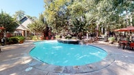 Located at 2330 Montgomery Park Blvd in Conroe, Texas, this outdoor amenity area offers a well-appointed resort-style pool environment nestled within a mature, wooded setting. The centerpiece is a generously sized freeform swimming pool with a smooth concrete surround, complemented by an attached spa feature along one edge. The pool's vivid turquoise water stands out against the natural backdrop of towering trees that provide substantial shade throughout the space.

The surrounding patio area is thoughtfully furnished for relaxation and socializing. Red-cushioned lounge chairs are arranged along the pool deck, offering comfortable spots for sunbathing or unwinding beneath the tree canopy. A shaded dining area with a red umbrella and matching table and chair sets provides an inviting space for outdoor gatherings. Lush tropical plantings and potted greenery are integrated throughout the deck, enhancing the natural, garden-like atmosphere.

The space is enclosed by a dark metal perimeter fence, adding a sense of privacy and security. Views of the surrounding multi-story brick residential buildings are visible through the trees, grounding the outdoor area within the broader community. The combination of dappled natural light, abundant greenery, and carefully selected furnishings gives this outdoor space a serene, retreat-like ambiance ideal for leisure and relaxation.