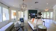This single-floor space showcases a thoughtfully designed open-concept layout that seamlessly blends a comfortable lounge area with a formal dining space. The living area is anchored by a crisp white sofa adorned with coral accent pillows, complemented by a dark-framed coffee table and a sleek black accent chair. A striking geometric pendant light serves as a central focal point, casting a warm, layered glow throughout the space.

A bold teal accent wall draws the eye toward the back of the room, where a wall-mounted flat-screen television is flanked by symmetrical sconce lighting, creating a polished media nook. The dining area transitions naturally from the lounge space, featuring a dark wood table surrounded by upholstered high-back chairs.

Natural light streams in through French doors along one wall, brightening the predominantly white and neutral palette. Recessed lighting supplements the natural illumination overhead. Lush indoor plants add organic texture and life to the corners of the room, while decorative ceramic vessels and styled accessories contribute to the space's refined, contemporary ambiance.