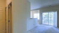 This single-floor space is designed with an open, airy feel, highlighted by an abundance of natural light streaming through multiple windows and a glass-paneled exterior door. The living area features wall-to-wall carpeting in a neutral tone, complementing the crisp, light-colored walls throughout. A defining architectural detail is the pass-through window connecting the kitchen to the living area, framed by a countertop ledge that serves as both a functional and visual divider between the two spaces. This design element enhances the open flow of the layout while maintaining a subtle separation between areas. The kitchen is positioned slightly elevated or set apart, with recessed or track lighting overhead adding focused illumination to the space. A glass-paneled door at the far end of the living area provides access to an outdoor balcony or patio, offering views of surrounding greenery and further enhancing the sense of openness. The overall ambiance is clean, bright, and neutral, providing a versatile foundation well-suited to a variety of interior styles and furnishing preferences.