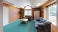 Located at 4909 E Upriver Dr in Spokane, Washington, this single-floor community room offers a welcoming and functional gathering space designed for both relaxation and casual dining. The interior is defined by warm terracotta and earthy orange walls that create an inviting atmosphere throughout, complemented by teal-green carpeting that extends across the entire floor.

The space is divided into two distinct functional zones. The lounge area features comfortable patterned sofas and armchairs arranged around a wooden coffee table, with a large wood armoire providing storage. A prominent arched window floods the seating area with natural light and adds an architectural focal point, while vaulted ceiling elements lend a sense of openness to this section.

The dining area accommodates multiple seating arrangements with several tall bistro-style tables and chairs suited for groups of varying sizes. Additional windows along the dining side provide views of the surrounding outdoor area and allow natural light to supplement the overhead fluorescent lighting. A textured ceiling adds subtle visual depth to this zone.

A private door at the rear of the space leads to an additional room or utility area. Decorative touches such as framed wall art and potted plants contribute to the comfortable, community-oriented character of the space overall.