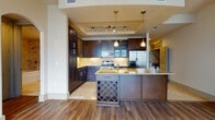 This single-floor high-rise residence offers a sophisticated urban living experience defined by sweeping city skyline views and refined interior finishes throughout. The open living area is anchored by rich hardwood flooring and expansive floor-to-ceiling glass walls that flood the space with natural light while framing dramatic views of the surrounding cityscape. Track lighting overhead adds a modern, functional touch.

The gourmet kitchen is thoughtfully designed with dark espresso cabinetry extending to the ceiling, stainless steel appliances including a French door refrigerator, electric range, and built-in microwave. Gold-toned granite countertops complement warm travertine tile flooring, while a large island features an integrated sink, additional seating, and a built-in wine rack beneath—ideal for entertaining.

The primary bathroom is a standout feature, offering a dual-sink vanity with dark cabinetry, a soaking tub clad in travertine tile, a glass-enclosed shower, and a window with city views. A secondary bathroom mirrors the design aesthetic with dark cabinetry, tile flooring, and framed mirror.

The bedroom features plush carpeting, large windows with city views, and sliding glass doors that open to a private brick-accented balcony, providing a seamless indoor-outdoor connection amidst the urban environment.