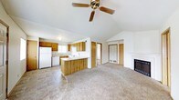 This single-floor home offers an inviting open-concept design that seamlessly connects the kitchen and living areas. The space is defined by striking vaulted ceilings that rise to a peak, creating a sense of airiness and volume, complemented by a ceiling fan that adds both style and function to the main living area. The kitchen is well-appointed with warm wood-toned cabinetry, white appliances including a refrigerator, dishwasher, and range, and a peninsula with a light-colored countertop that provides additional workspace and casual seating. Tile flooring in the kitchen transitions naturally into plush carpeting throughout the living area, adding warmth underfoot. The living room is anchored by a classic fireplace, serving as a focal point for the space. Natural light filters in through the kitchen window, brightening the heart of the home. Multiple doorways lead to additional rooms beyond, suggesting a practical and well-organized floor plan. The clean, neutral color palette throughout creates a versatile backdrop, making the space adaptable for a variety of interior styles. Overall, this home combines comfortable living with practical design on a single, easy-to-navigate level.