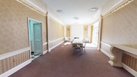 This single-floor space showcases a blend of traditional and refined architectural elements throughout its interior. The main room is generously proportioned, offering an open and airy feel enhanced by large windows along one wall that flood the space with natural light. Recessed circular ceiling fixtures provide additional illumination, complementing the bright white ceiling and crisp crown molding that frame the room elegantly.

The walls feature a warm, textured treatment in soft golden-beige tones, divided by white wainscoting panels and pilasters that lend a formal, classical character to the space. A fireplace serves as a focal point at one end of the room, flanked by white accent chairs that add a touch of understated sophistication.

A dining or conference table with chairs is positioned centrally, making the space well-suited for meetings, gatherings, or events. The dark, patterned carpeting grounds the room and contrasts with the lighter wall and ceiling tones. An adjacent doorway leads to a secondary area with a distinctive tiled floor, visible through louvered doors, suggesting additional functional space nearby. Overall, the atmosphere is formal yet welcoming.