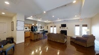 This single-floor space offers an expansive open-concept design that seamlessly connects the kitchen, dining, and living areas into one cohesive environment. The vaulted ceilings rise dramatically overhead, creating a sense of generous vertical space and an airy atmosphere throughout. Recessed lighting and pendant fixtures provide well-distributed illumination, complementing the natural light that streams in through large windows and French doors along the back wall.

The kitchen is equipped with wooden cabinetry, a black refrigerator, and a center island with bar-height seating, making it both functional and social. The island serves as a natural transition point between the kitchen and the open dining and living spaces beyond.

Rich, dark hardwood floors extend throughout the entire area, adding warmth and visual continuity. The living area features a comfortable seating arrangement anchored by a fireplace, above which a wall-mounted television is positioned. Ceiling fans are installed throughout to support climate comfort. French doors and side windows invite natural light and provide outdoor access, enhancing the overall openness of the space.
