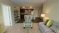 This single-floor residence offers a thoughtfully designed open-concept layout that connects the living area and kitchen in one cohesive, flowing space. The living room is anchored by plush carpeting and features ample room for comfortable seating arrangements, complemented by a decorative open shelving unit that adds both storage and visual interest.

The kitchen is a standout feature, showcasing rich dark cabinetry that extends to the ceiling, stainless steel appliances including a refrigerator, microwave, and range, and a central island with a light stone countertop. Industrial-style pendant lights hang above the island, providing focused task lighting while enhancing the modern aesthetic. Bar-height stools line the island, creating a casual dining and entertaining space.

Hardwood-style flooring flows throughout the kitchen and adjoining areas, providing a clean contrast to the carpeted living space. Neutral wall tones create a warm and inviting ambiance, while the open sightlines between rooms make the overall space feel bright and expansive. A hallway visible in the background suggests additional private rooms beyond the main living area. The overall design balances functionality with contemporary style, making it well-suited for everyday living and entertaining alike.