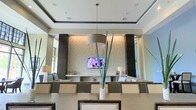Situated at 211 River Landing Dr. on Daniel Island, South Carolina, this refined single-floor space presents a seamlessly designed interior that blends modern aesthetics with comfortable functionality. The main gathering area commands attention with its tray ceilings accented by dark trim details, lending a sense of architectural depth and verticality to the room. Recessed lighting is integrated throughout, providing consistent, even illumination across the space.

A striking textured stone accent wall serves as a central focal point, flanked by sleek dark vertical columns that frame a built-in media display. Open shelving on either side of the wall provides both a decorative and functional element, styled with tasteful accessories and accent lighting.

The dining area features a large communal table surrounded by nailhead-trim upholstered chairs, anchored by a substantial drum pendant light overhead. A tall sculptural plant arrangement adds a natural, organic contrast to the otherwise contemporary palette of neutral creams, whites, and dark espresso tones.

The open layout allows for a clear sightline across the space, suggesting a fluid connection between the dining and lounge areas. Overall, the space exudes a polished, upscale character well-suited for social gatherings and community engagement.